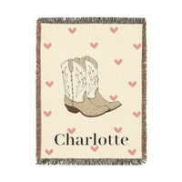 Personalized woven baby blanket with cowgirl boots and soft pink hearts on cream, with a name woven in, shown flat