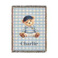 Personalized woven baby blanket with a teddy bear in a blue cap and sweater on light blue gingham, with a name woven in, shown flat