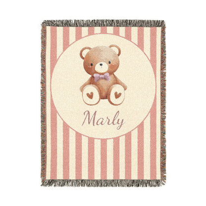 Personalized woven baby blanket with a brown teddy bear in a purple bow tie and little hearts in a cream circle on a blush pink and cream stripe, with a name woven in, shown flat