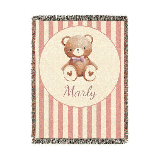 Personalized woven baby blanket with a brown teddy bear in a purple bow tie and little hearts in a cream circle on a blush pink and cream stripe, with a name woven in, shown flat