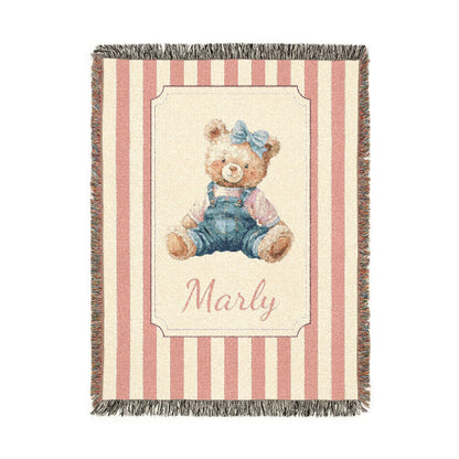 Personalized woven baby blanket with a watercolour teddy bear in dungarees and a blue bow on a pink and cream stripe, with a name woven in, shown flat