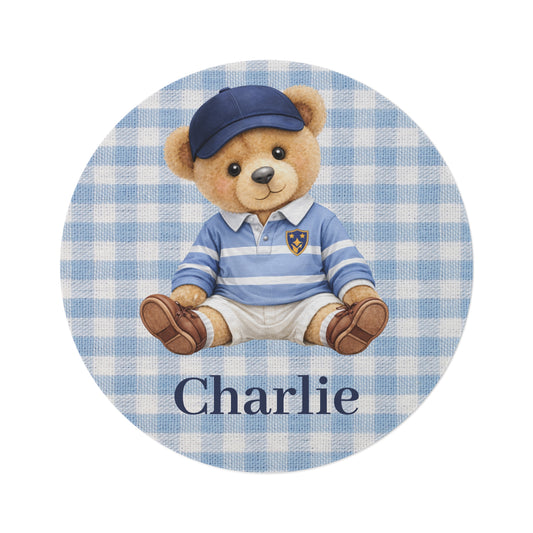 Personalized round nursery rug with a teddy bear in a blue cap and striped rugby shirt on blue gingham, with a name woven in, shown flat