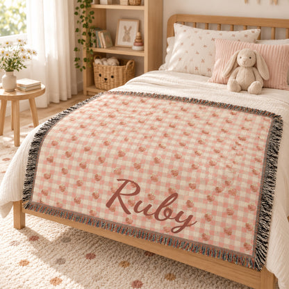 Personalized strawberry gingham woven blanket laid across a toddler bed in a soft nursery, name Ruby at the foot of the bed