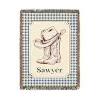 Personalized woven baby blanket with a cowboy hat and boots on a cream panel with a blue gingham border and a name woven in, shown flat