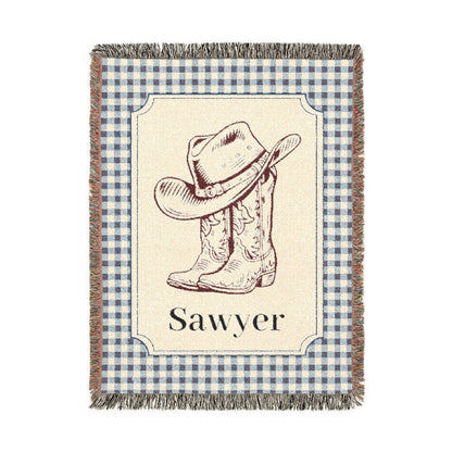 Personalized woven baby blanket with a cowboy hat and boots on a cream panel with a blue gingham border and a name woven in, shown flat
