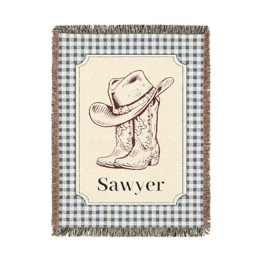 Personalized woven baby blanket with a cowboy hat and boots on a cream panel with a blue gingham border and a name woven in, shown flat