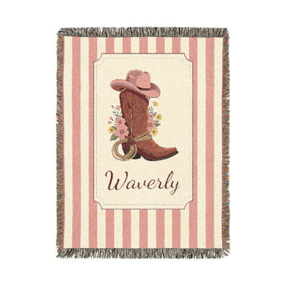 Personalized woven baby blanket with a cowgirl boot, pink hat and wildflowers in a framed panel on a pink and cream stripe, with a name woven in, shown flat