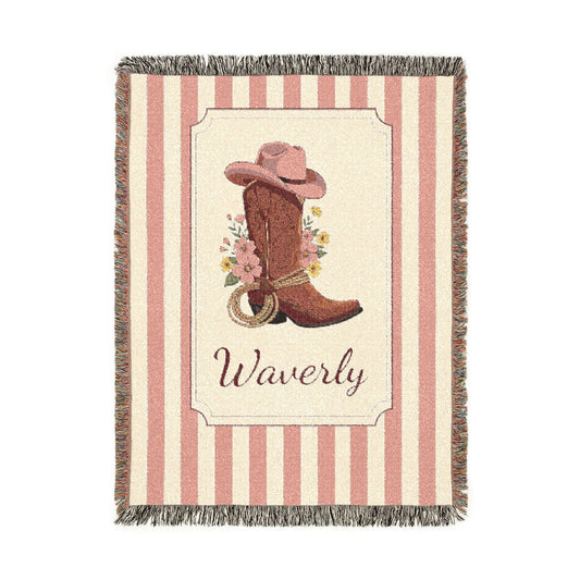 Personalized woven baby blanket with a cowgirl boot, pink hat and wildflowers in a framed panel on a pink and cream stripe, with a name woven in, shown flat