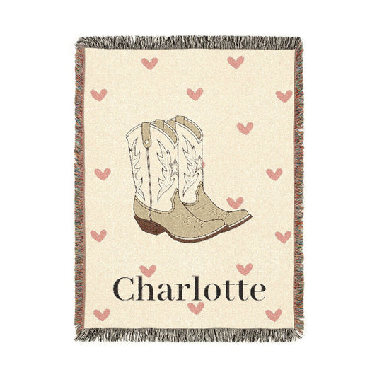 Personalized woven baby blanket with cowgirl boots and soft pink hearts on cream, with a name woven in, shown flat