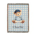 Personalized woven baby blanket with a teddy bear in a blue cap and sweater on light blue gingham, with a name woven in, shown flat