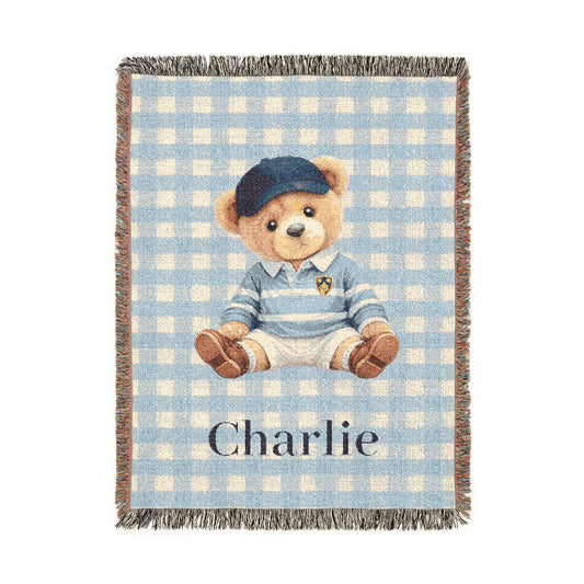 Personalized woven baby blanket with a teddy bear in a blue cap and sweater on light blue gingham, with a name woven in, shown flat